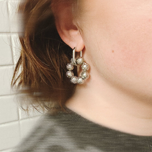 Close-up of an ear wearing a silver hoop earring with clear stones against a tiled wall background.