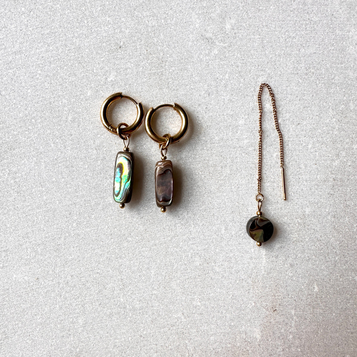 A pair of gold-toned hoop earrings with rectangular abalone shell pendants, displayed next to a matching long drop earring with a faceted dark bead on a textured light gray background.