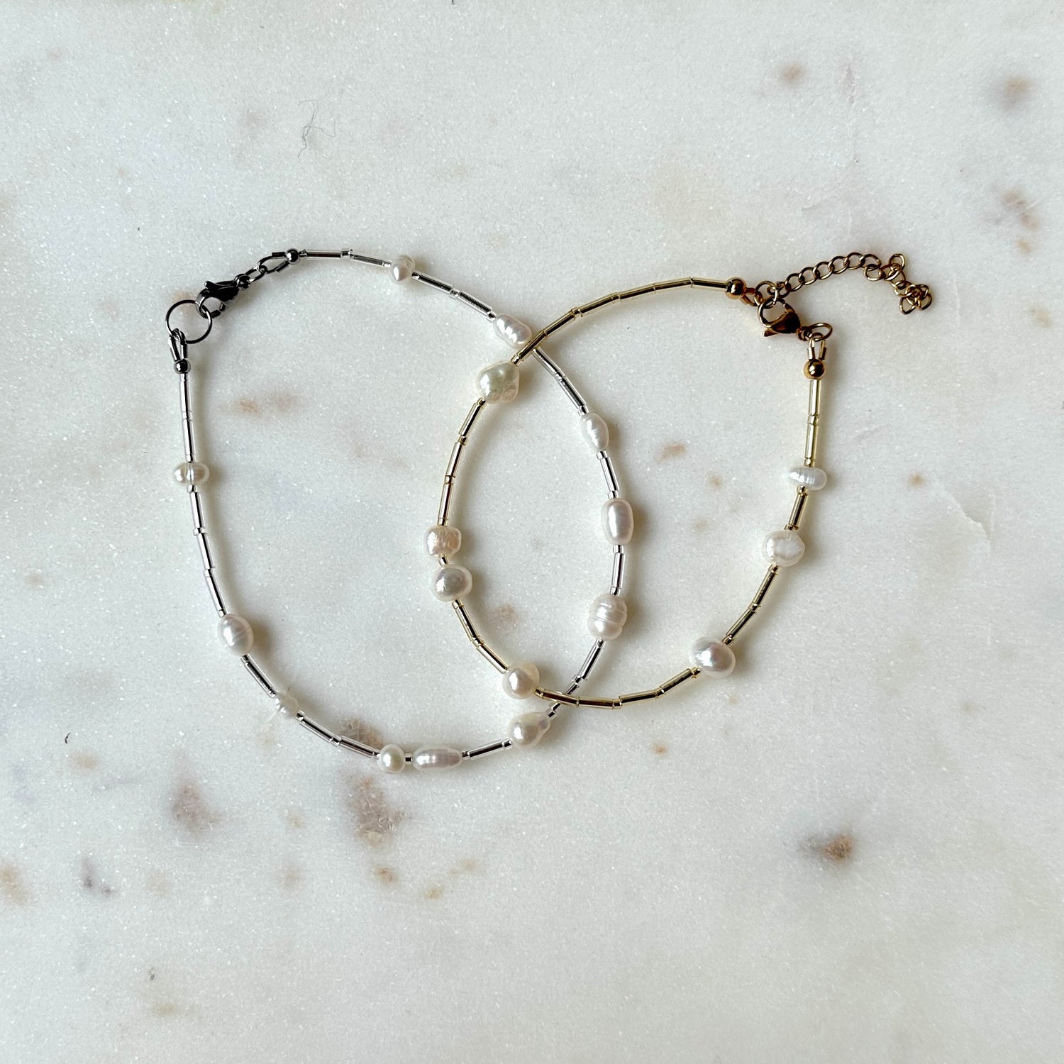 Two pearl and glass bead anklets on a stainless steel wire, laid out on a marble surface.
