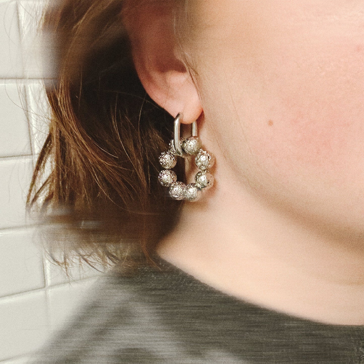 Close-up of an ear wearing a silver hoop earring with clear stones against a tiled wall background.