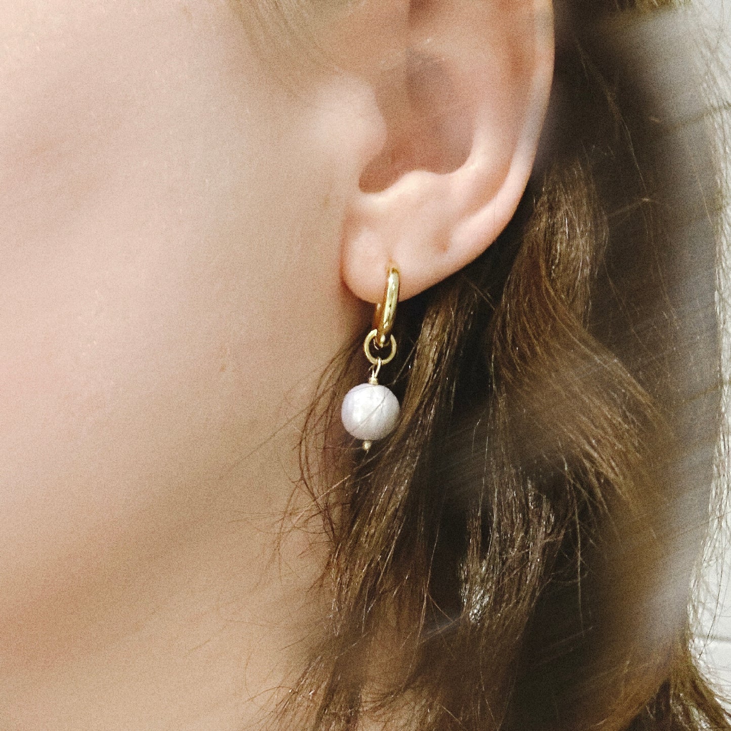 Gold earring with a white blue drop worn by a person with brown hair.