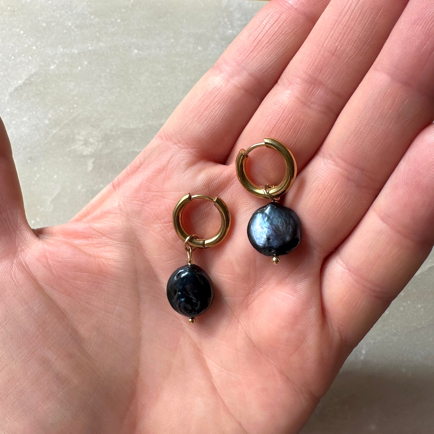 Gold hoop earrings with black pearls held in a hand against a neutral background