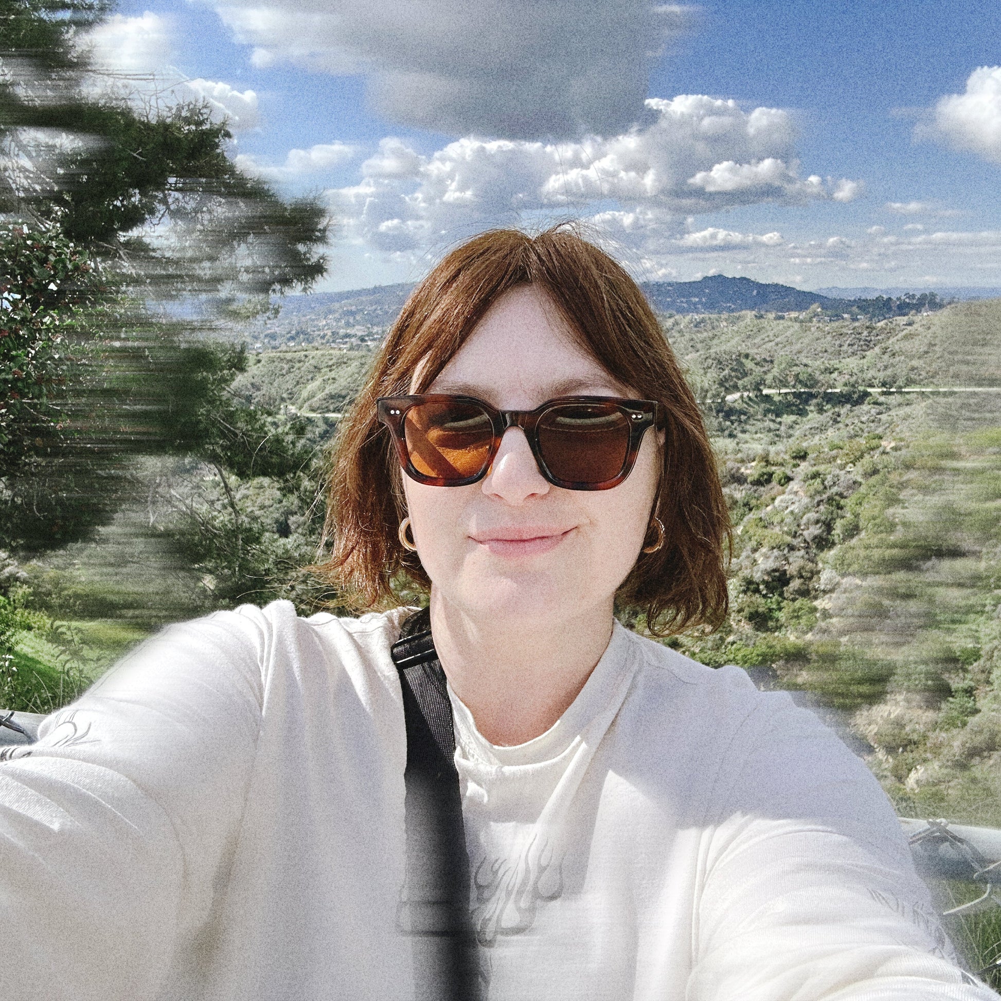 Person wearing sunglasses and gold chunky earrings with a scenic background of Los Angeles and the surrounding mountains.