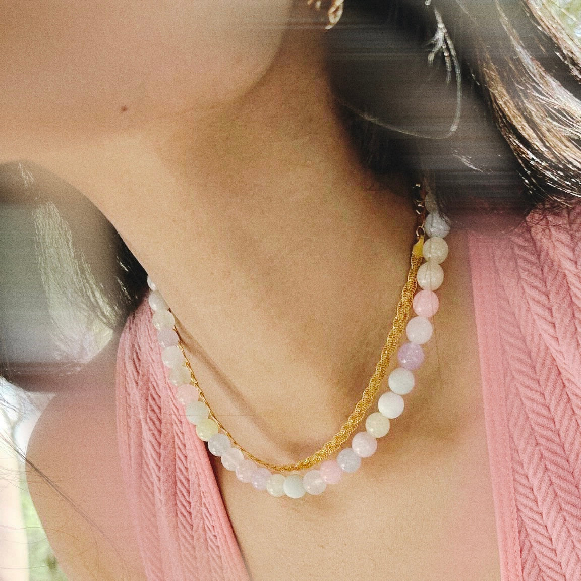 Close-up of a person wearing a handmade colourful stone necklace, a gold necklace and a pink top with blurred background